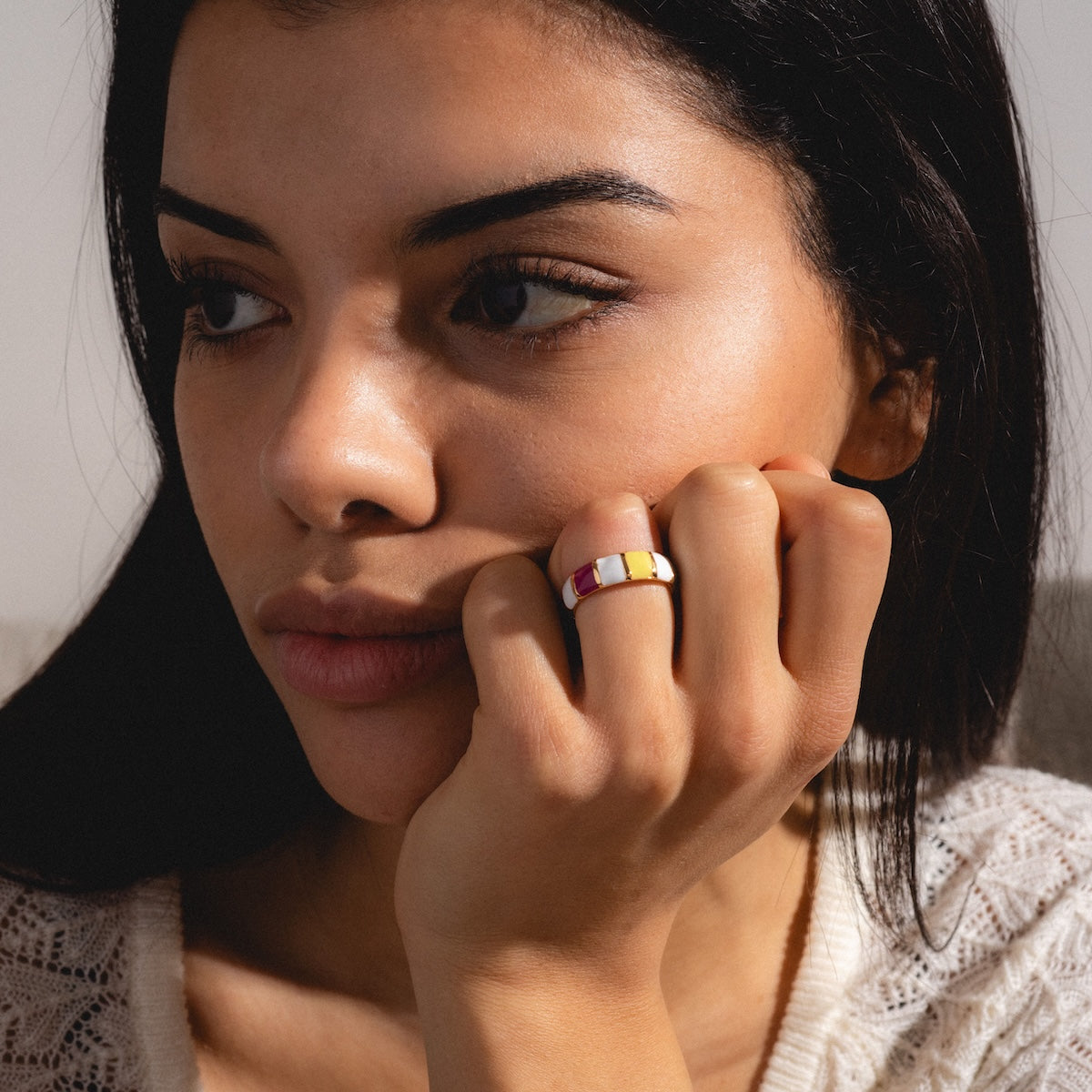 A woman with long dark hair and a thoughtful expression rests her chin on her hand, showcasing the Chroma Ring—a 24k gold plated ring featuring red, white, and yellow segments—while wearing a cream knit sweater.