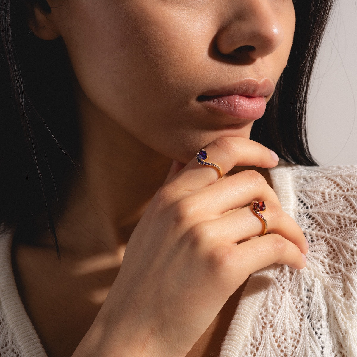 A woman with long dark hair touches her chin, showcasing the Main Character Floating Ombre Birthstone Ring alongside another gold gemstone piece. She wears a white textured top, bathed in soft sunlight that highlights her smooth skin and natural makeup.