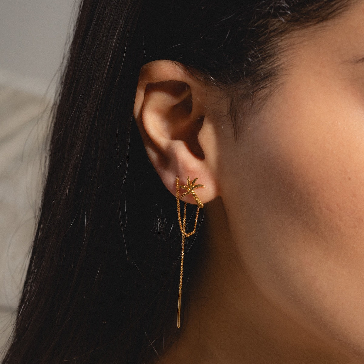 A close-up of a person’s ear wearing the Isla Earrings—24k gold plated with a starburst design and thin dangling chains. The person has long, dark hair and smooth skin.