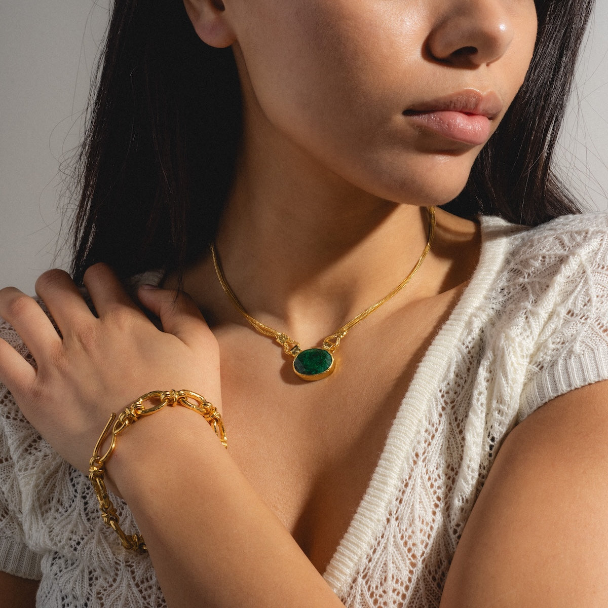 A woman in a white knit top showcases the Zehra Chain Bracelet and a gold necklace with a green gemstone pendant. Her hand, adorned with the Zehra bracelet, rests on her shoulder, partly covering the necklace.