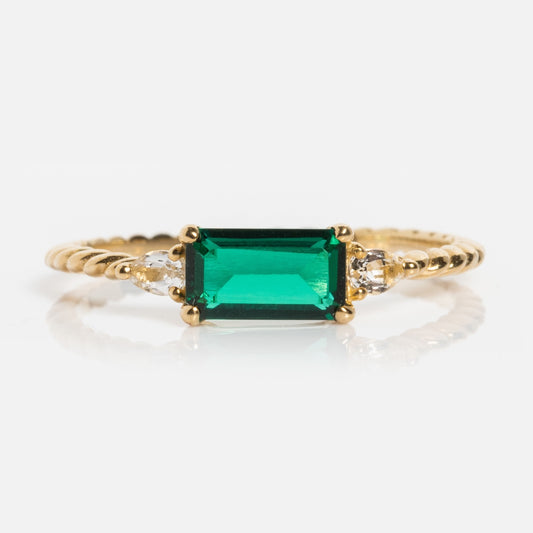 The Solid Gold 2025 May Capsule Classic Emerald Statement Ring features a twisted band, a rectangular green gemstone centerpiece, and two small round clear stones, shown against a white background.