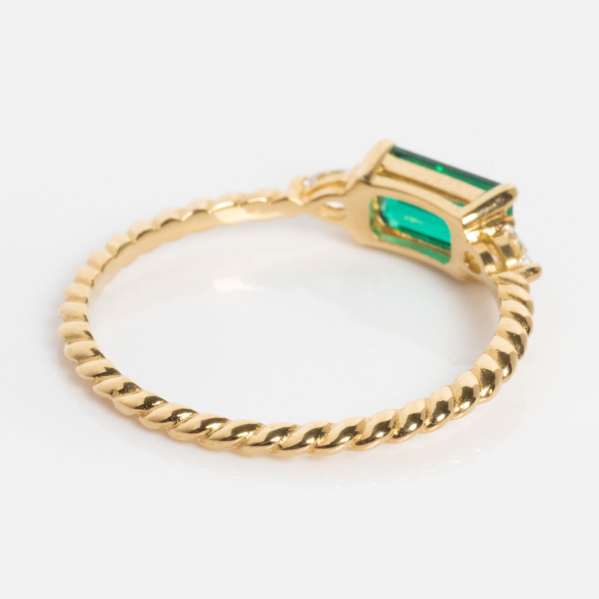 The Solid Gold 2025 May Capsule Classic Emerald Statement Ring features a twisted gold band and a rectangular green gemstone in a prong setting, creating a dazzling statement against a white background.