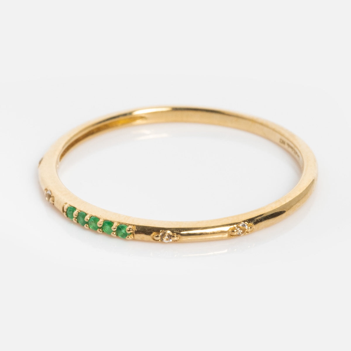 The Solid Gold 2025 May Capsule Delicate Emerald Stacking Ring showcases a slim gold band with a row of small green emeralds and a few tiny clear stones, set against a plain white background.