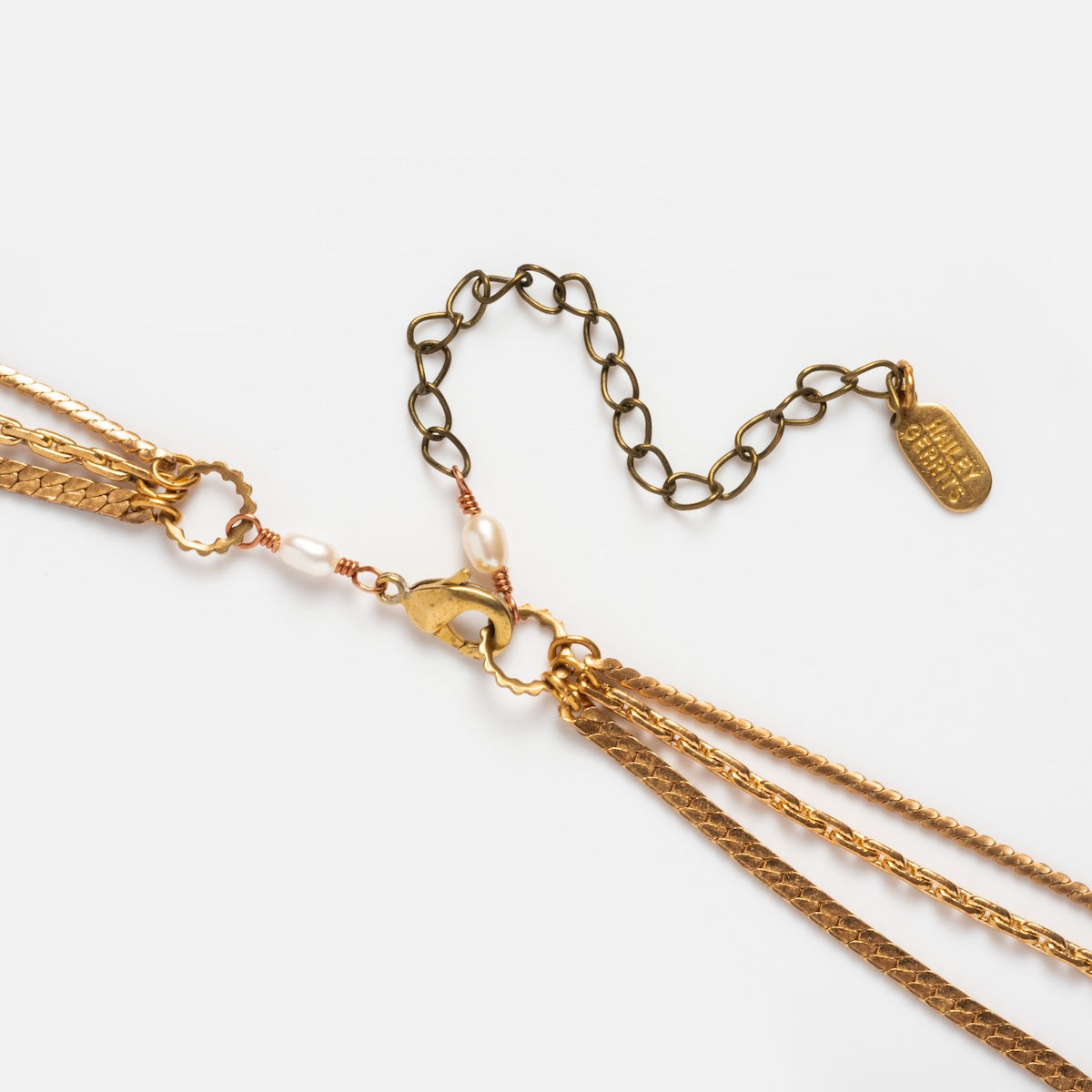 Close-up of the Palmer Gemstone Necklace: a gold layered chain with pearls, recycled brass chains, a lobster clasp, and an adjustable chain with a small rectangular engraved tag. Shown against a white background.