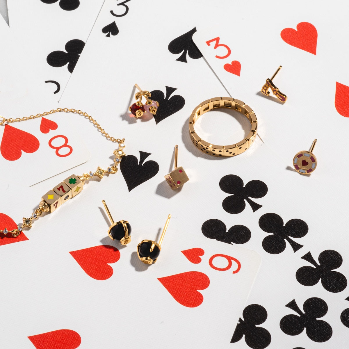 Magic 8 Ball Studs and gold-plated jewelry with playing card motifs are displayed on scattered cards featuring hearts, clubs, and spades.