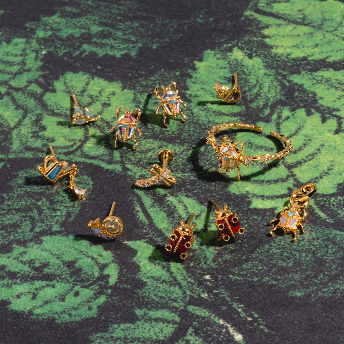 The Dragonfly Flat Back Stud, a gold vermeil earring with CZ and colorful gemstone accents, is displayed on a green leafy patterned background.