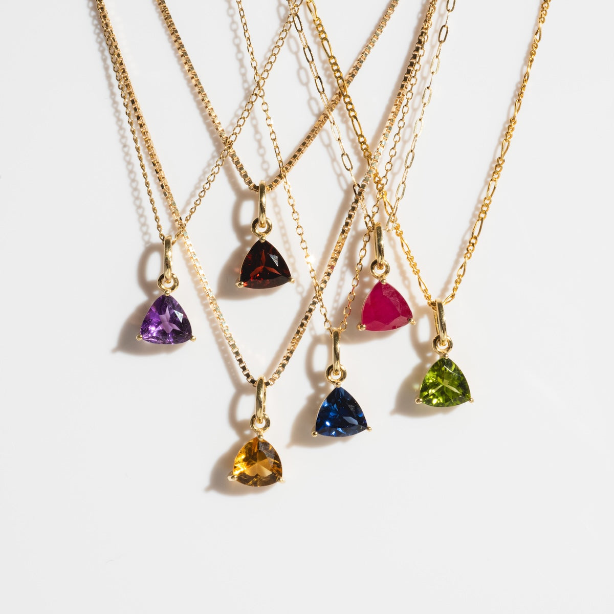 Six Solid Gold Trillion Birthstone Charm necklaces in purple, red, pink, blue, yellow, and green are displayed on a white background with their gold chains artfully overlapping.