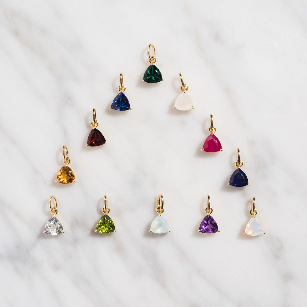 Twelve Family Gold Solid Gold Trillion Birthstone Charms with vibrant gemstones are displayed in a semicircle on a white marble surface.