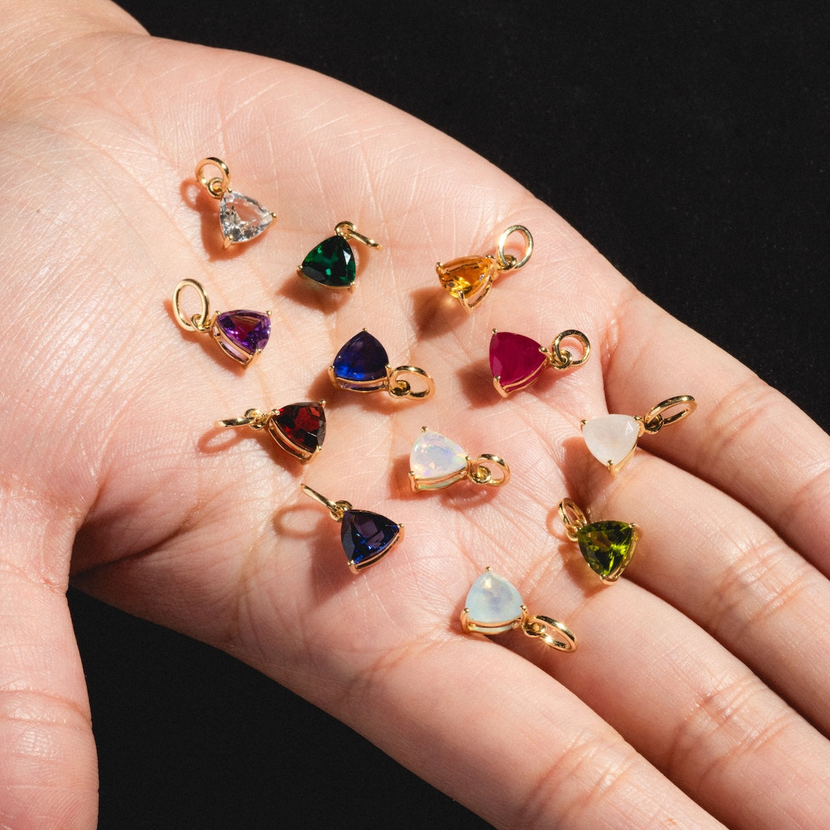 A hand displays Solid Gold Trillion Birthstone Charms in vibrant red, green, blue, yellow, white, and purple hues, each with a small gold frame and trillion cut stones, set against a black background.