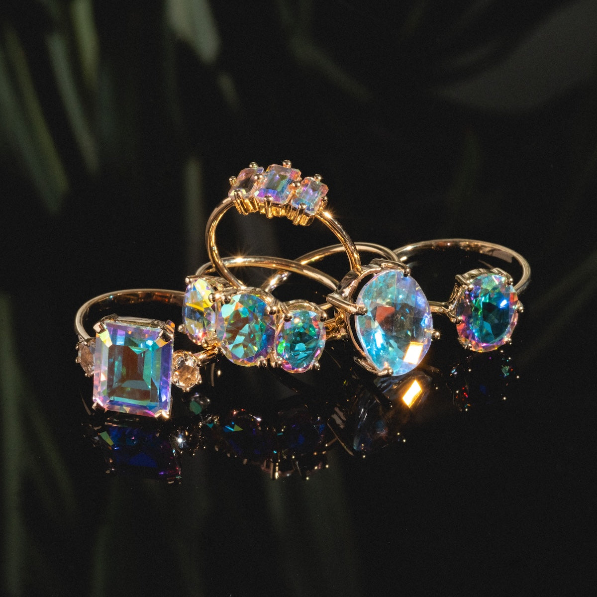Five gold rings, including the One of a Kind 14k Large Emerald Cut Angel Aura Ring, are displayed on shiny black. Their iridescent gemstones shimmer in blue, purple, and pink hues, casting colorful reflections around them.