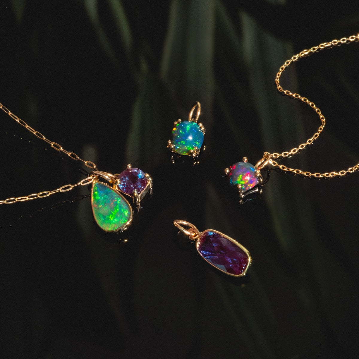 The One of a Kind 14k Elongated Pear Drop Black Welo Opal Charm is displayed on a glossy black surface with a subtle leafy background, highlighting its unique teardrop shape and vibrant gemstone for a standout jewelry look.