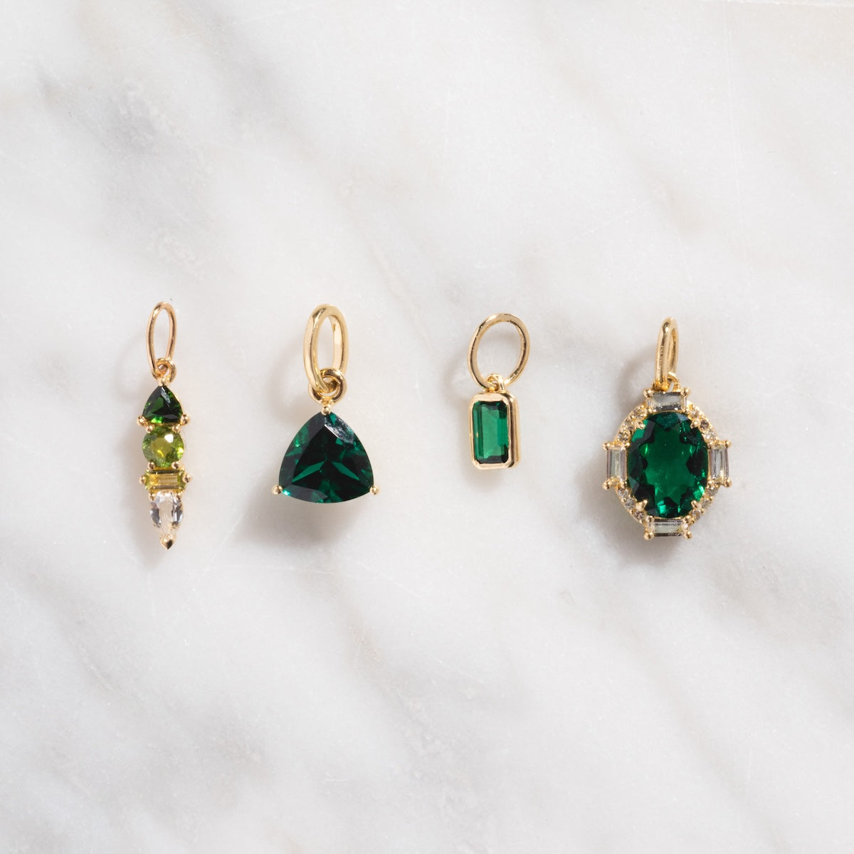 Four 9k yellow gold pendants with green gemstones—teardrop, triangle, rectangle, and oval—are lined up on white marble. The Solid Gold Trillion Birthstone Charm stands out with its eye-catching trillion-cut stone.