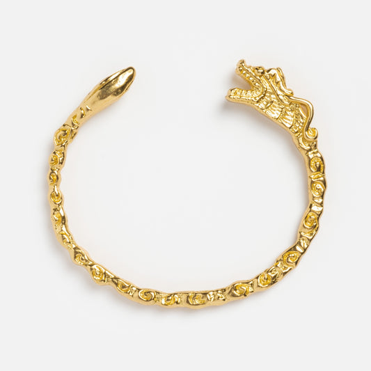 The Auspicious Bracelet is a gold dragon-shaped piece featuring a detailed dragon head on one end and a pointed tip on the other, displaying an intricate dragon motif against a plain white background.
