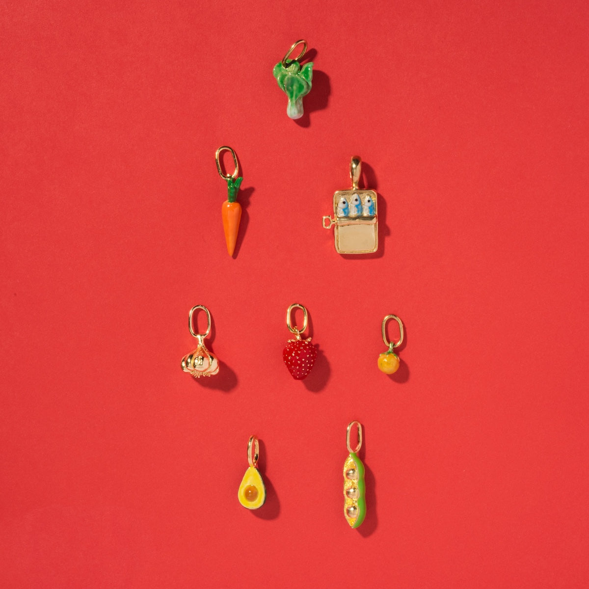 Eight playful enamel Charms by Satomi Studio—broccoli, carrot, milk carton, garlic, strawberry, orange, avocado, and peas—are neatly arranged in rows on a vibrant red background.