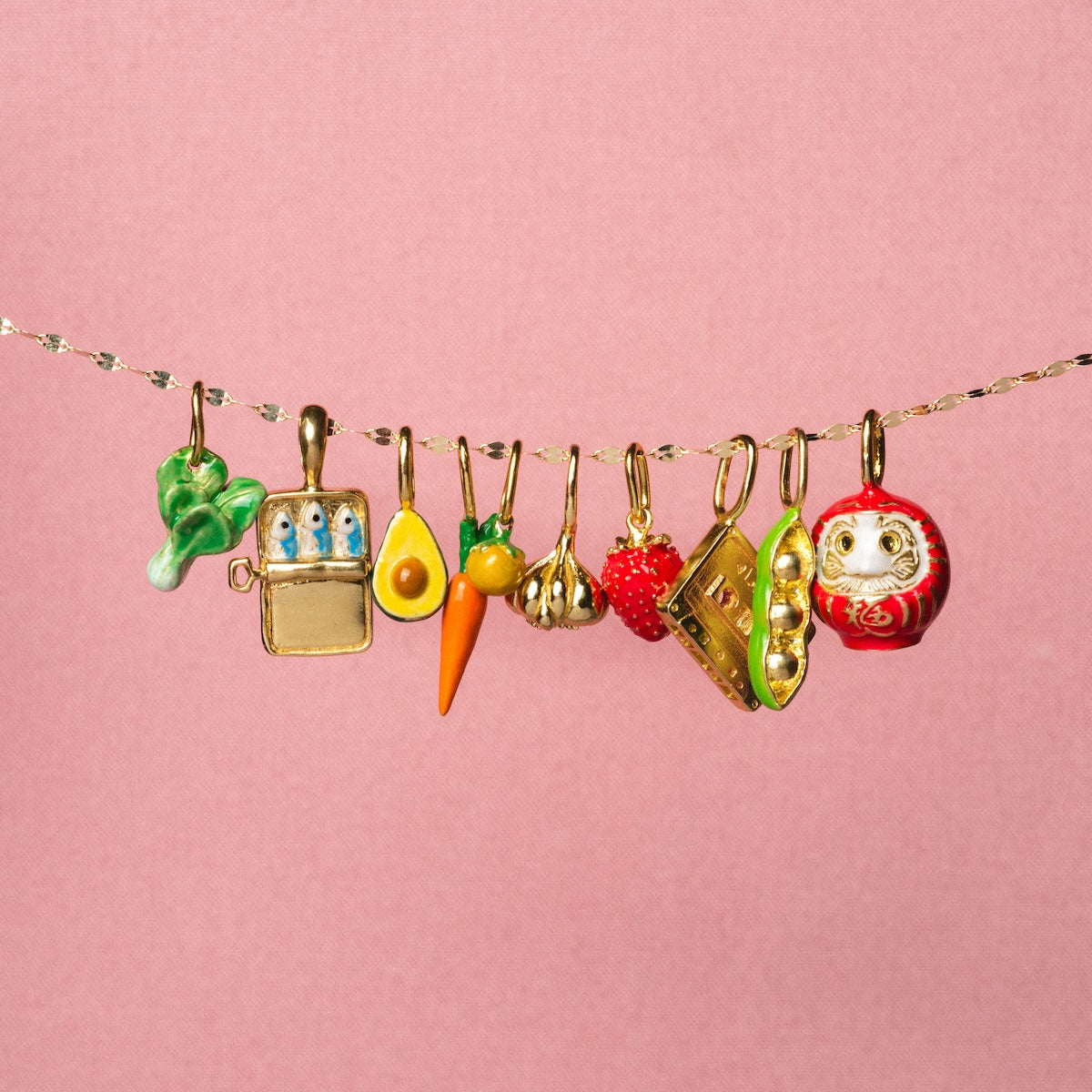 A row of nine playful Charms by Satomi Studio hangs from a gold chain against a pink background, featuring colorful enamel shapes like broccoli, paint palette, egg, and carrot—perfect as unique necklace pendants.