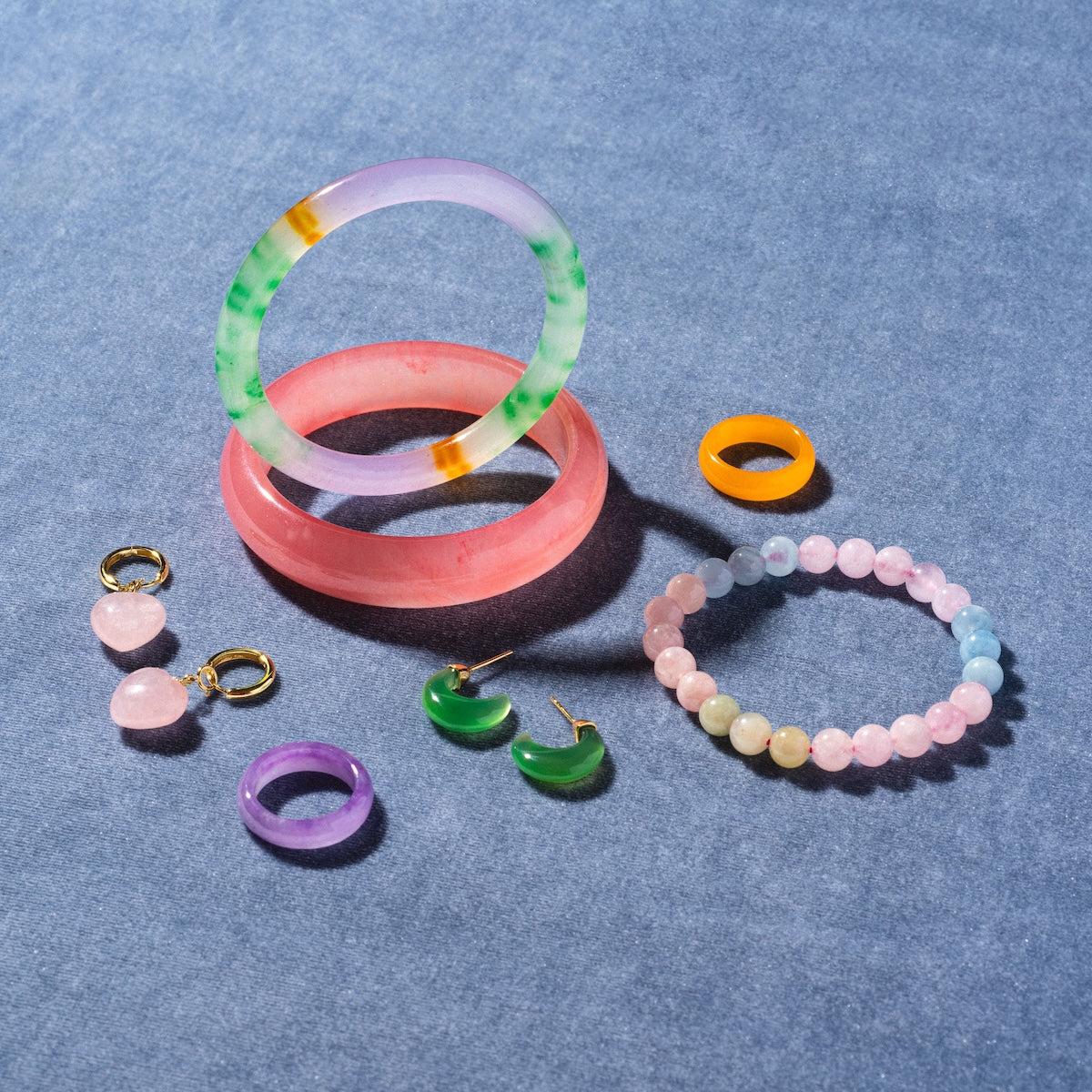 A collection of colorful jewelry on a blue surface, featuring 18k gold plated Half Moon Green Chalcedony Earrings, two bangles, a beaded bracelet, purple hoops, and an orange ring.