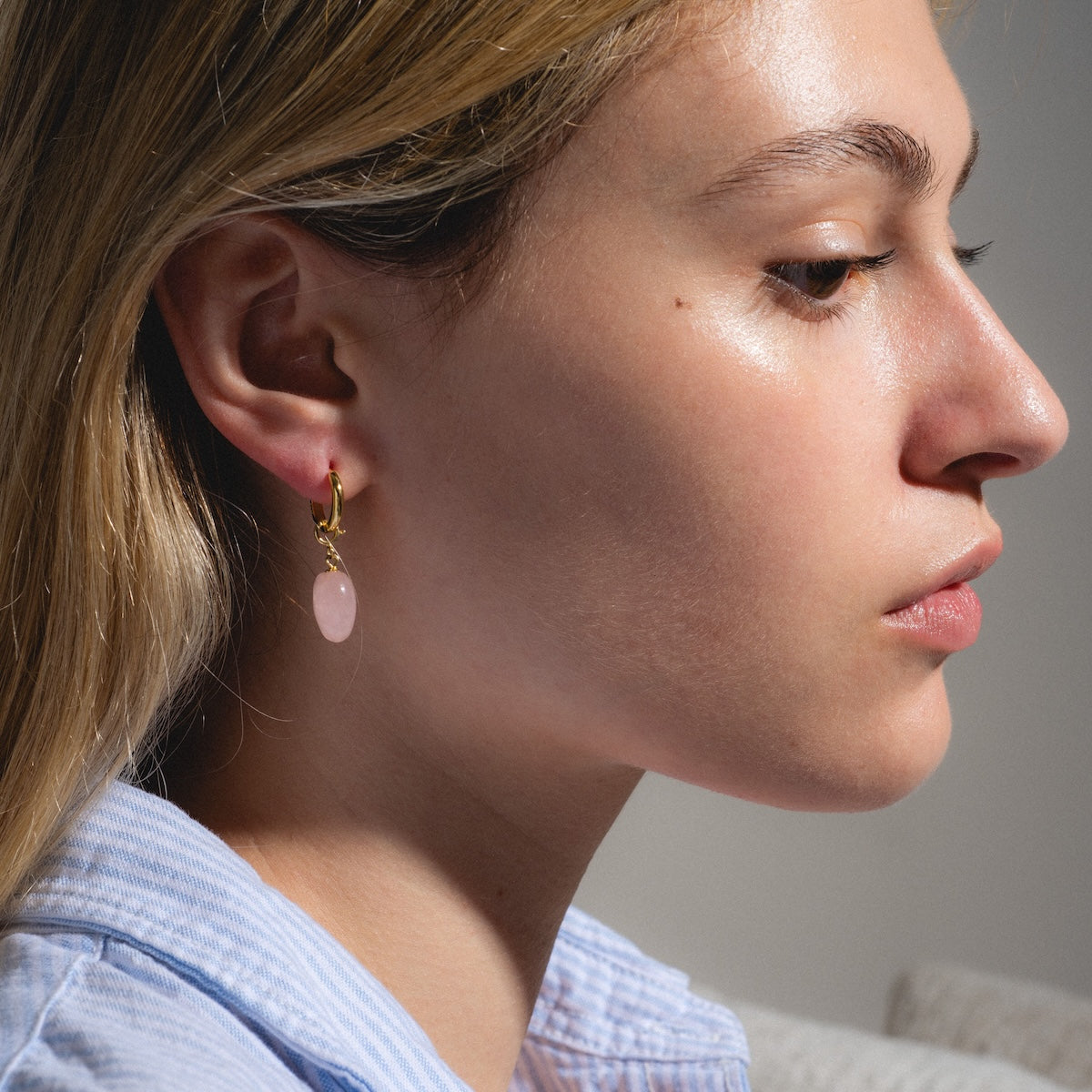 A close-up side profile of a woman with light skin and blonde hair wears a light blue shirt and Quartzite Heart Earrings—gold huggie hoops featuring a pink stone pendant—captured in natural light.