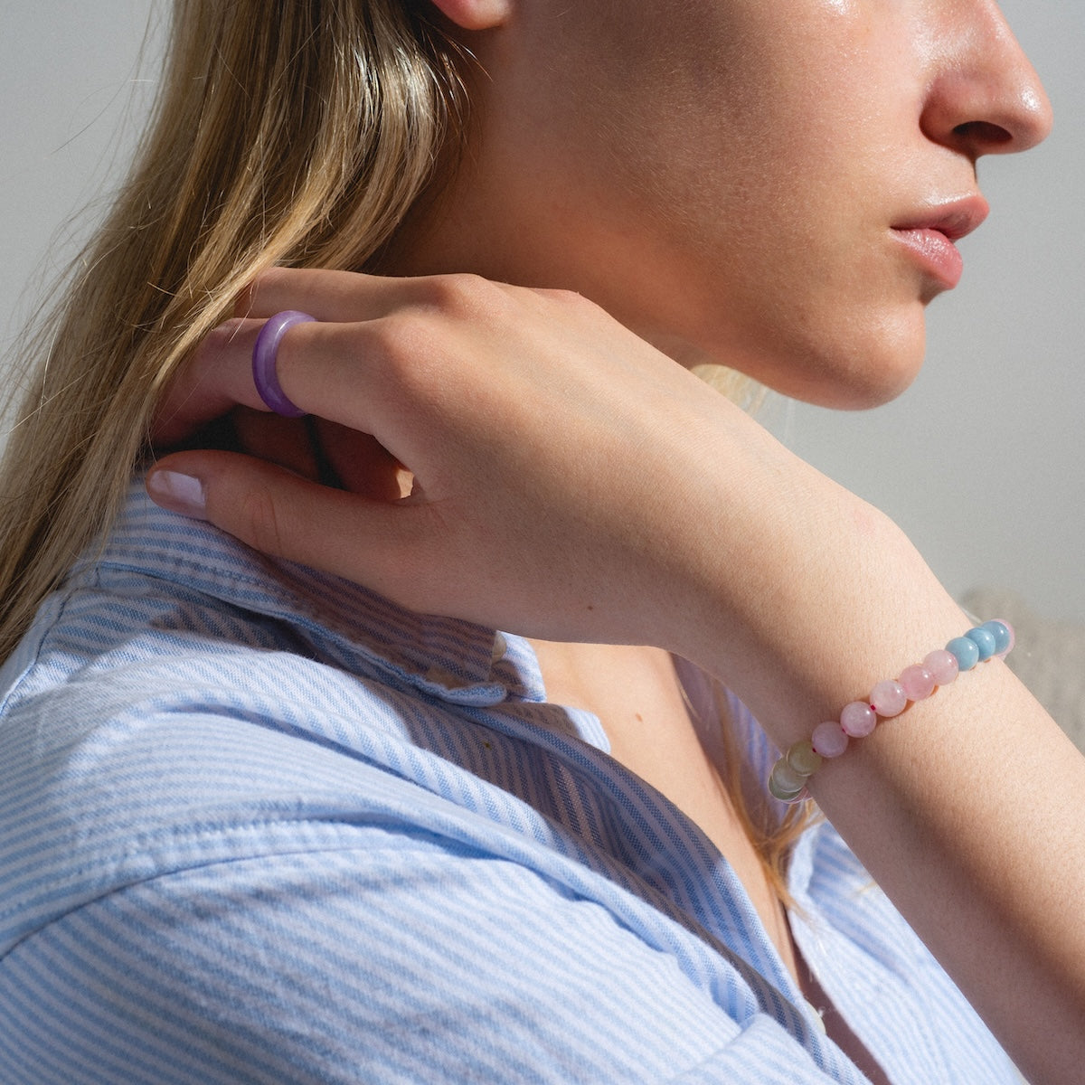 A woman in a blue and white striped shirt touches her neck, sunlight highlighting her face and hand as she wears the Billie Beaded Crystal Bracelet and a translucent purple ring.