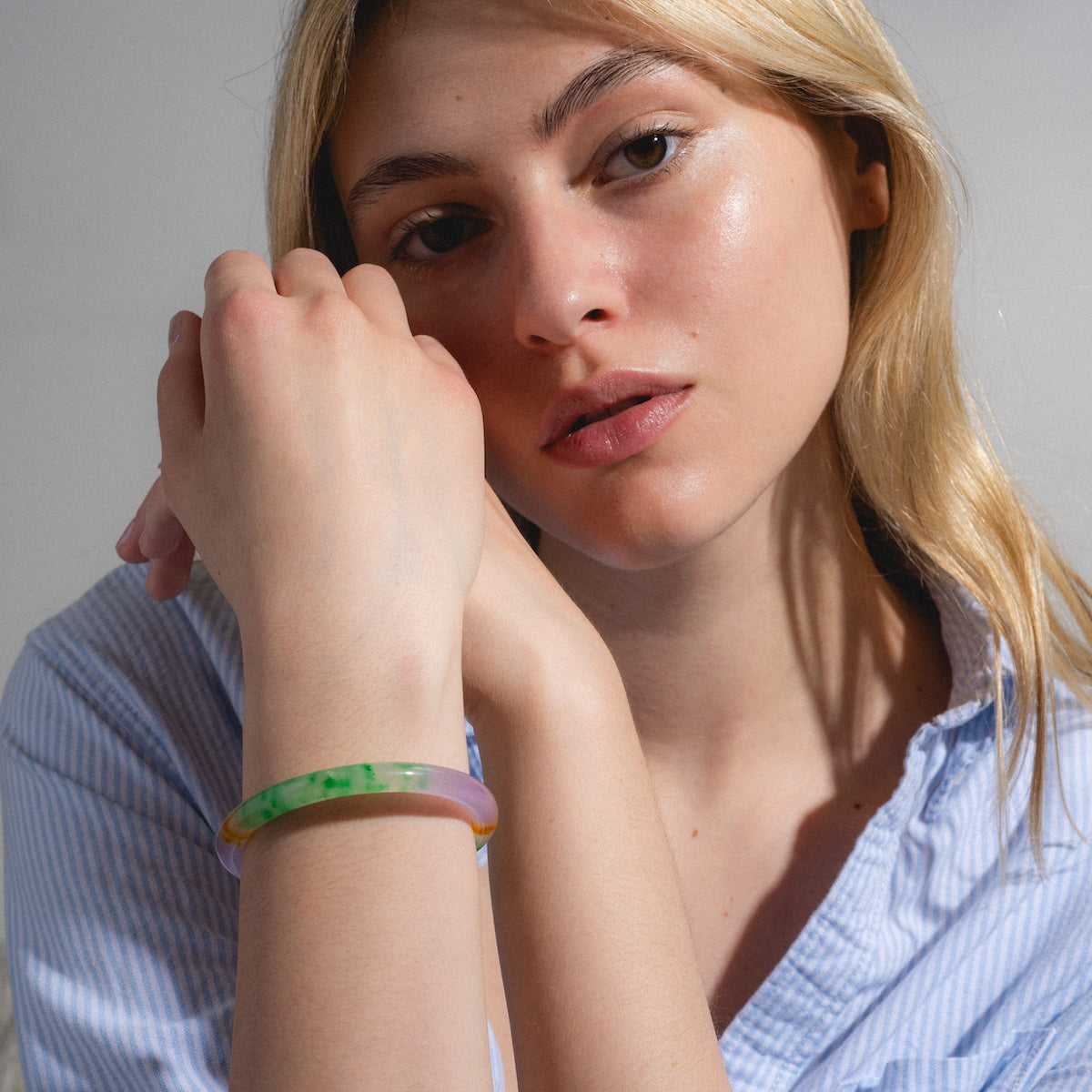 A young blonde woman in a light blue striped shirt rests her hand on her chin, thoughtfully showcasing the Lavender Field Multicolor Quartzite Bangle against a neutral background and highlighting its artistic design.