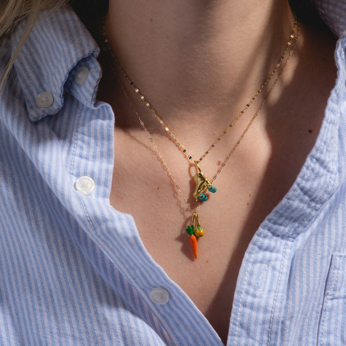 Close-up of a person wearing a light blue and white striped shirt, showcasing two Charms by Satomi Studio gold necklaces—one with playful blue enamel charms and the other with orange carrot and green pea brass charms.