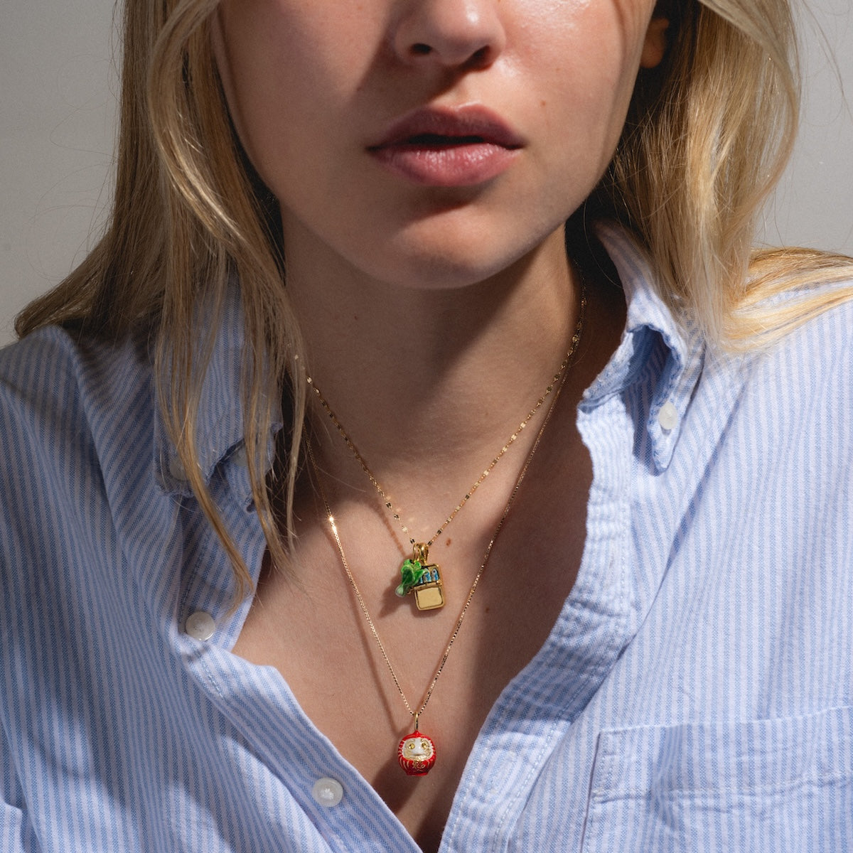 A woman wears a light blue, striped button-down shirt and two gold Charms by Satomi Studio necklaces—one with enamel pineapple and juice box charms, the other with a red and white round brass charm. Her face is partially visible.