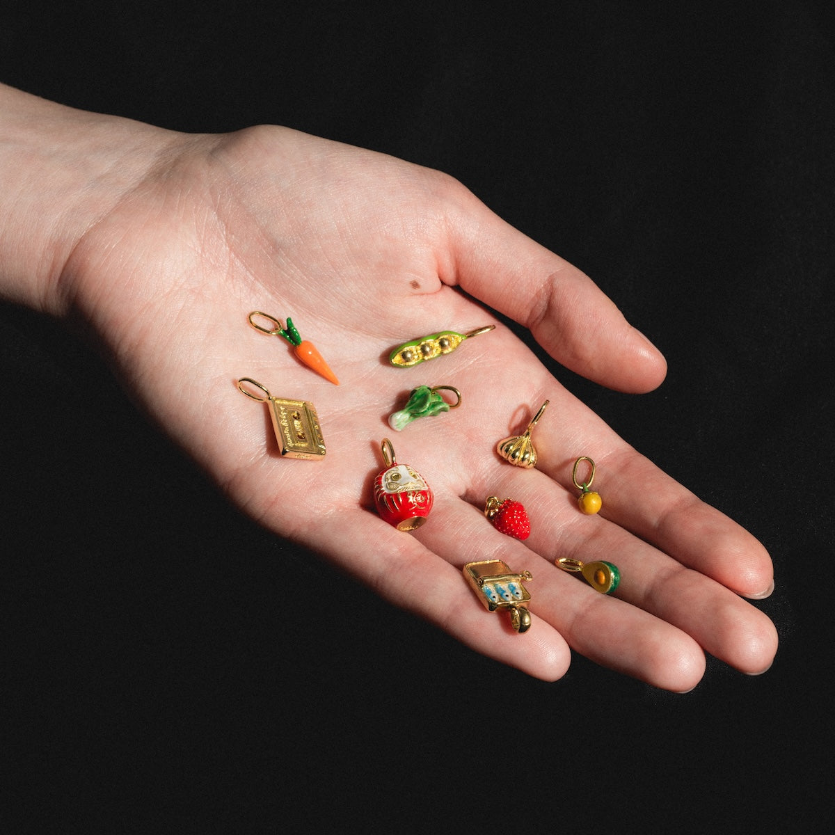 A hand with palm up displays ten colorful Charms by Satomi Studio, including a carrot, peas in a pod, a book, a red figurine, and a strawberry—ideal for playful enamel jewelry or adding to brass charm collections.