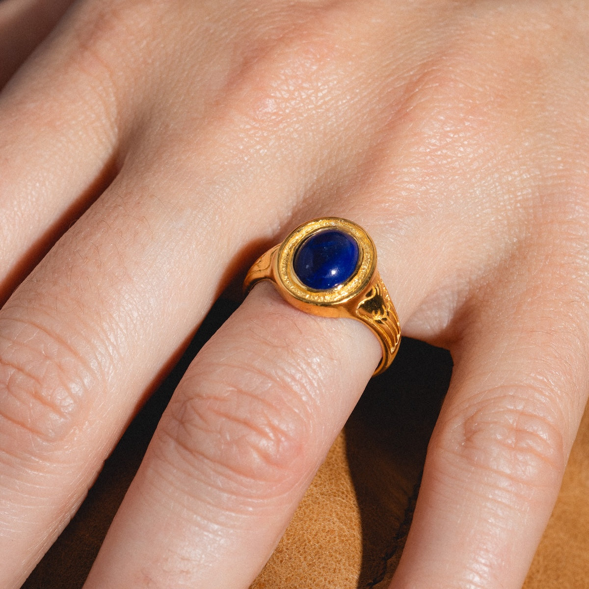 A hand wears the Astral Ring, featuring an oval dark blue Lapis stone set in gold vermeil, resting on a tan leather surface.
