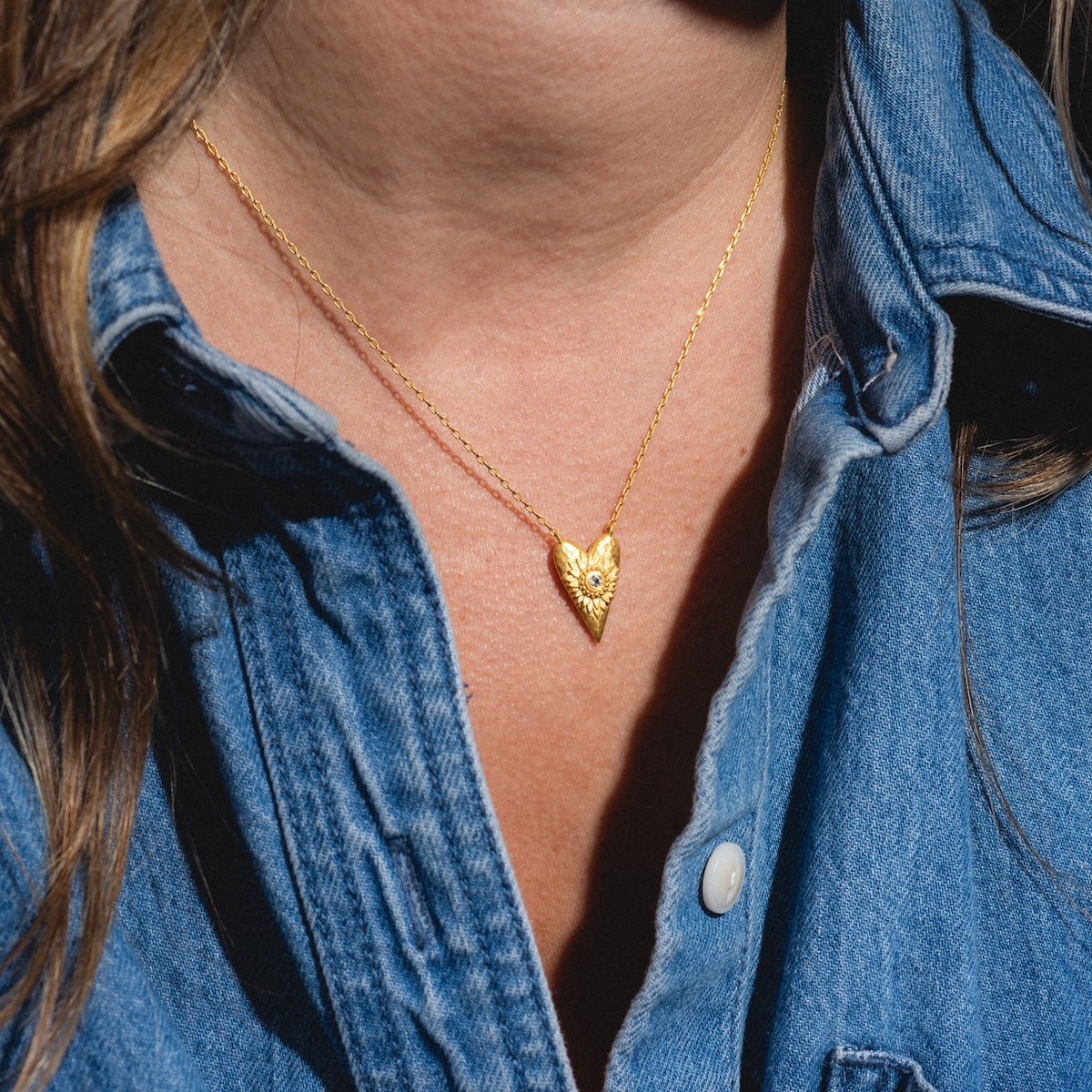 A person wearing a blue denim shirt and the I Am Loved Heart Necklace, which features a gold heart pendant with a detailed sunburst design.