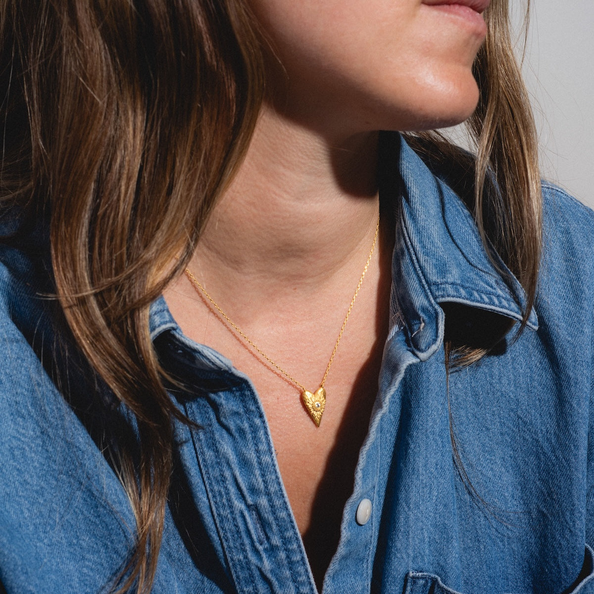 A woman wearing a blue denim shirt and the I Am Loved Heart Necklace, featuring gold and white topaz, shown from the shoulders up and facing slightly to the side.