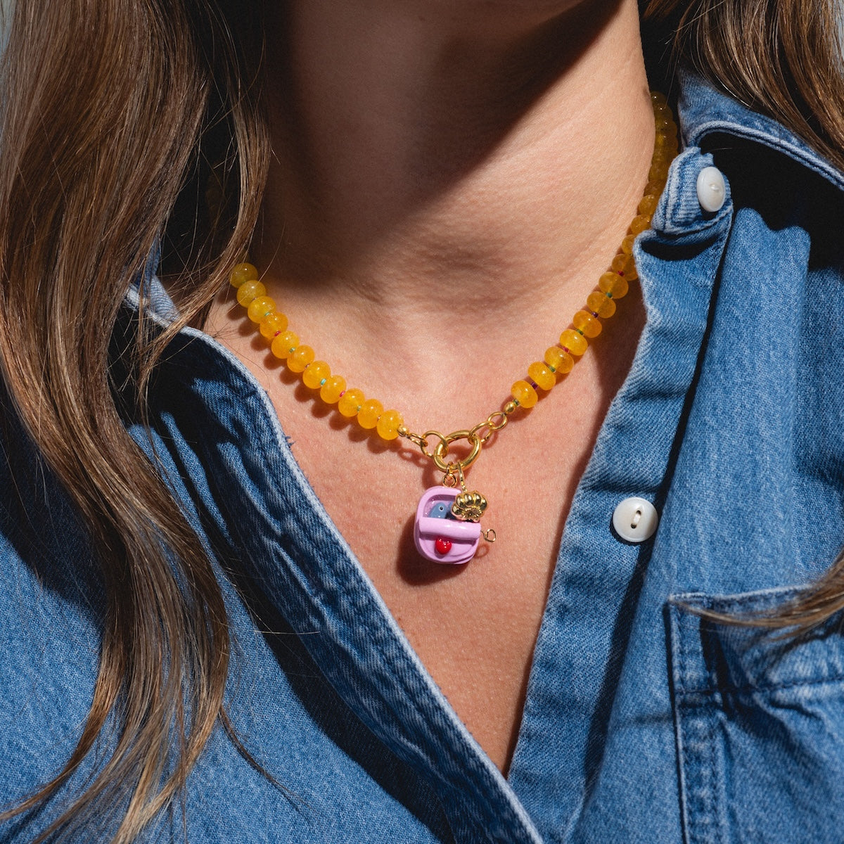 A person wears a blue denim shirt and the Charms by Warm Clay Co yellow beaded necklace, featuring hand-crafted, gold-plated clay charms like a pink, purple, and gold mouth-and-burger charm.