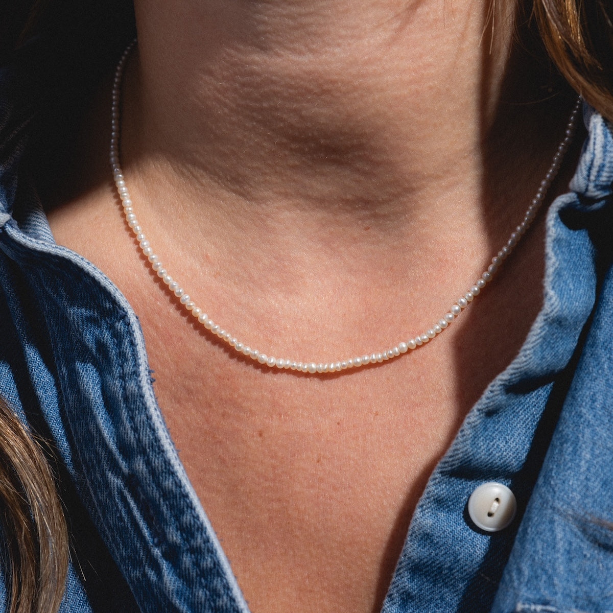 A close-up of a person wearing the Vermeil Delicate Pearl Chain with a blue denim shirt featuring white buttons, shown from neck to upper chest.
