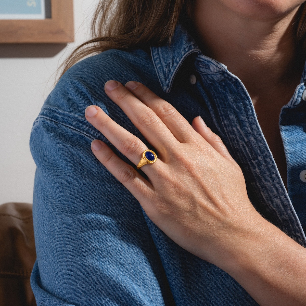 Wearing a blue denim shirt, a person rests their hand on their shoulder, showcasing the Astral Ring—a gold vermeil piece set with a round blue stone that shimmers in the bright light.