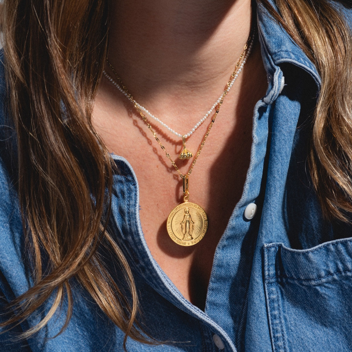 A person wears a blue denim shirt and layered gold and pearl necklaces, including the Chain For Charms with a bold medallion. Among the layers is a trendy paperclip chain, and the persons hair is partially visible.