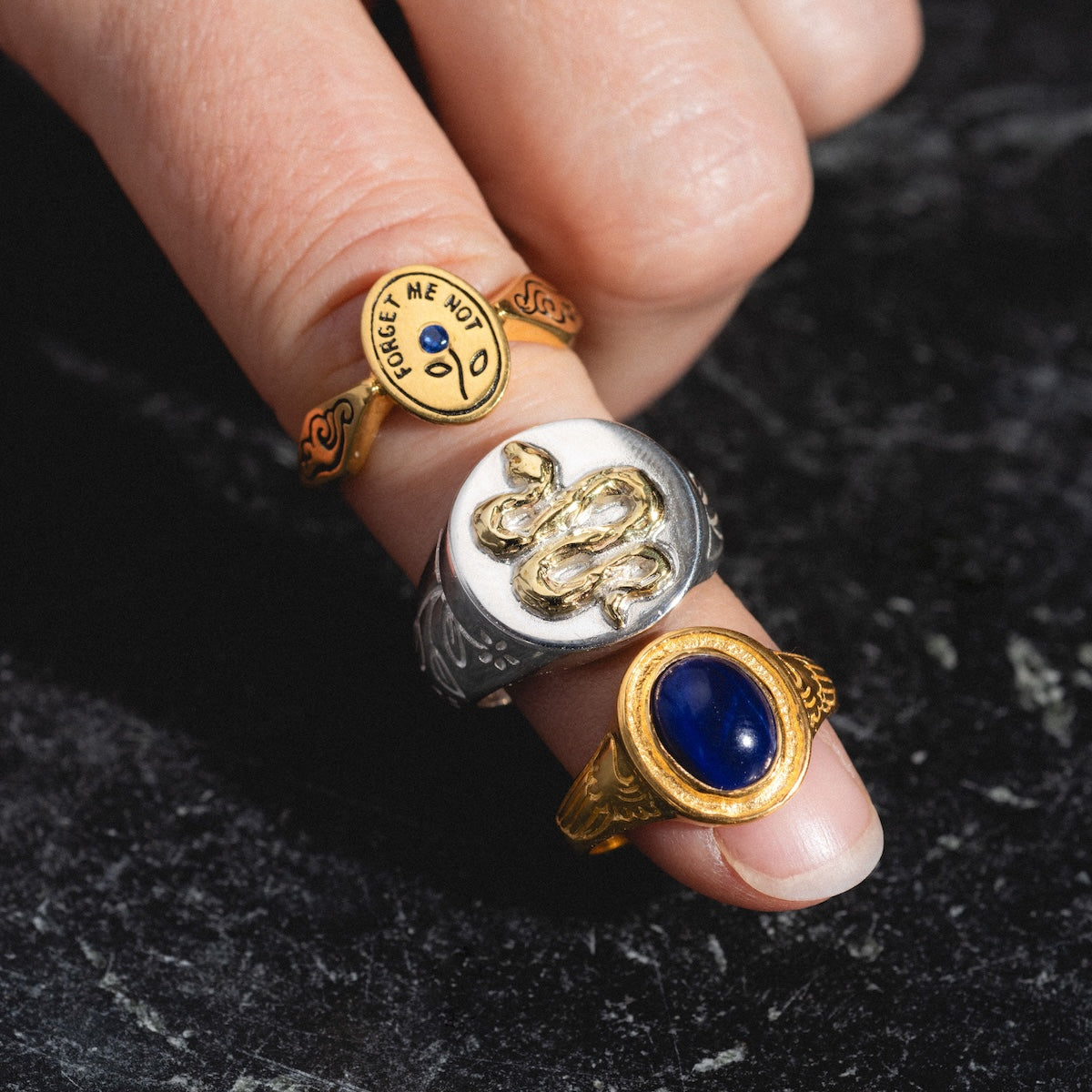 A hand adorned with three unique rings—one is the gold vermeil Astral Ring with a face, the second a silver ring featuring a raised snake design, and the third a lapis ring with an oval dark blue stone—all set against a dark, textured background.