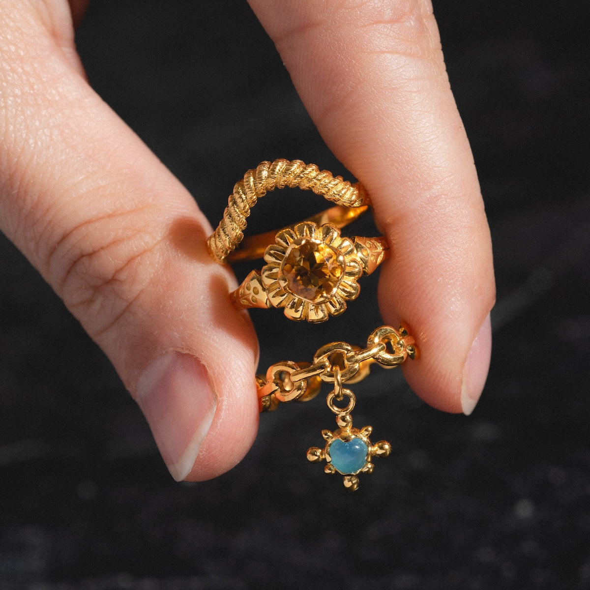 A hand with neatly trimmed nails holds two rings—an 18k gold vermeil sunflower ring and the Curved Rope Ring, a stacking style featuring a small blue gemstone charm—shown against a dark background.