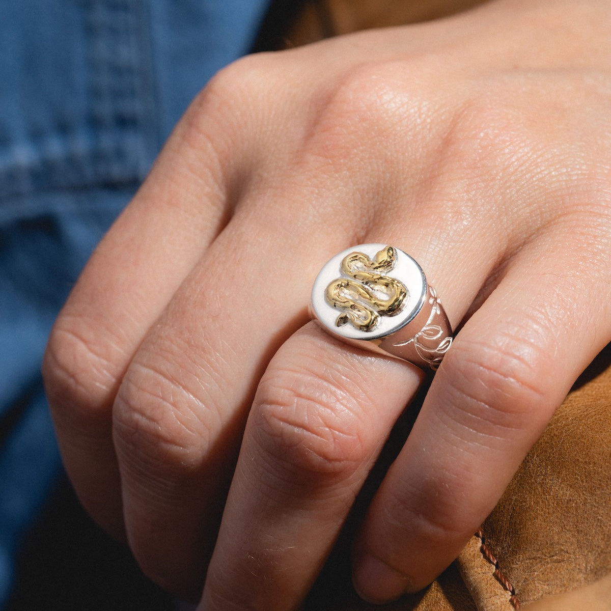A close-up of a hand wearing the Serpent Signet Ring, featuring a raised gold vermeil serpent on sterling silver. The wearer holds a light brown leather item and has on a blue denim garment.