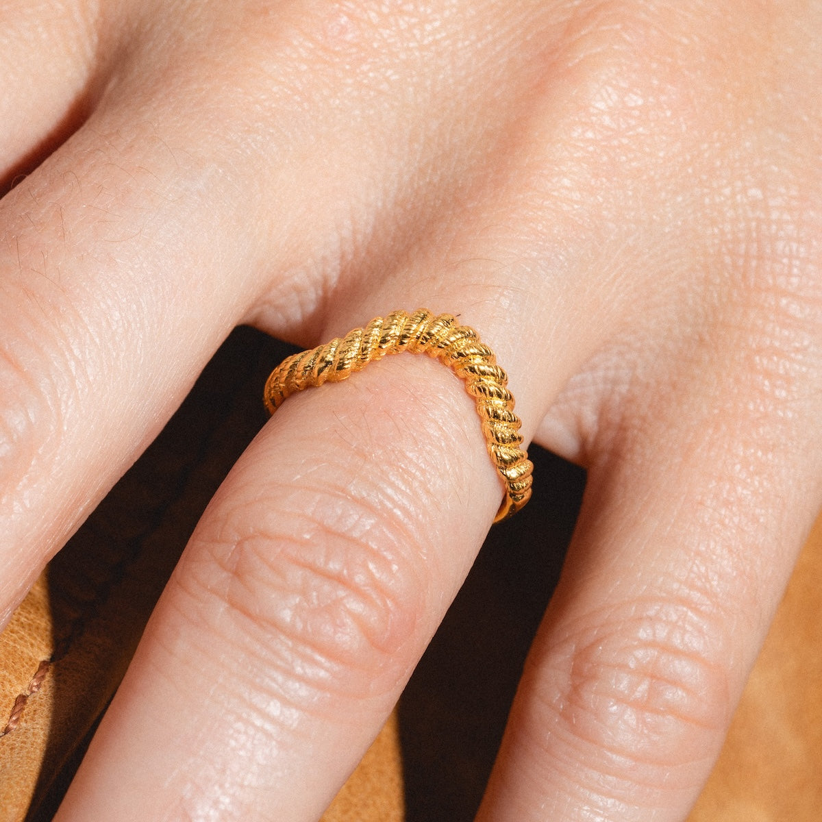 A close-up of a hand wearing the Curved Rope Ring, crafted in 18k gold vermeil, on the middle finger. The light-toned skin contrasts softly against a brown fabric background.