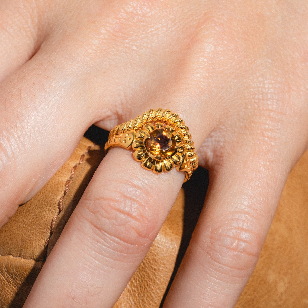 A close-up of a hand wearing the Curved Rope Ring—an 18k gold vermeil piece with a sunflower-inspired rope design and a round amber gemstone center—resting on brown leather.