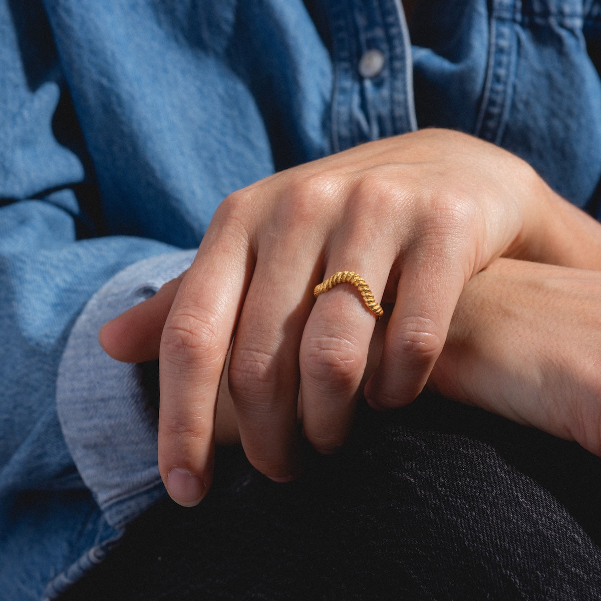 A hand wearing the Curved Rope Ring in 18k gold vermeil, styled with a blue denim shirt and dark pants, rests on a knee.