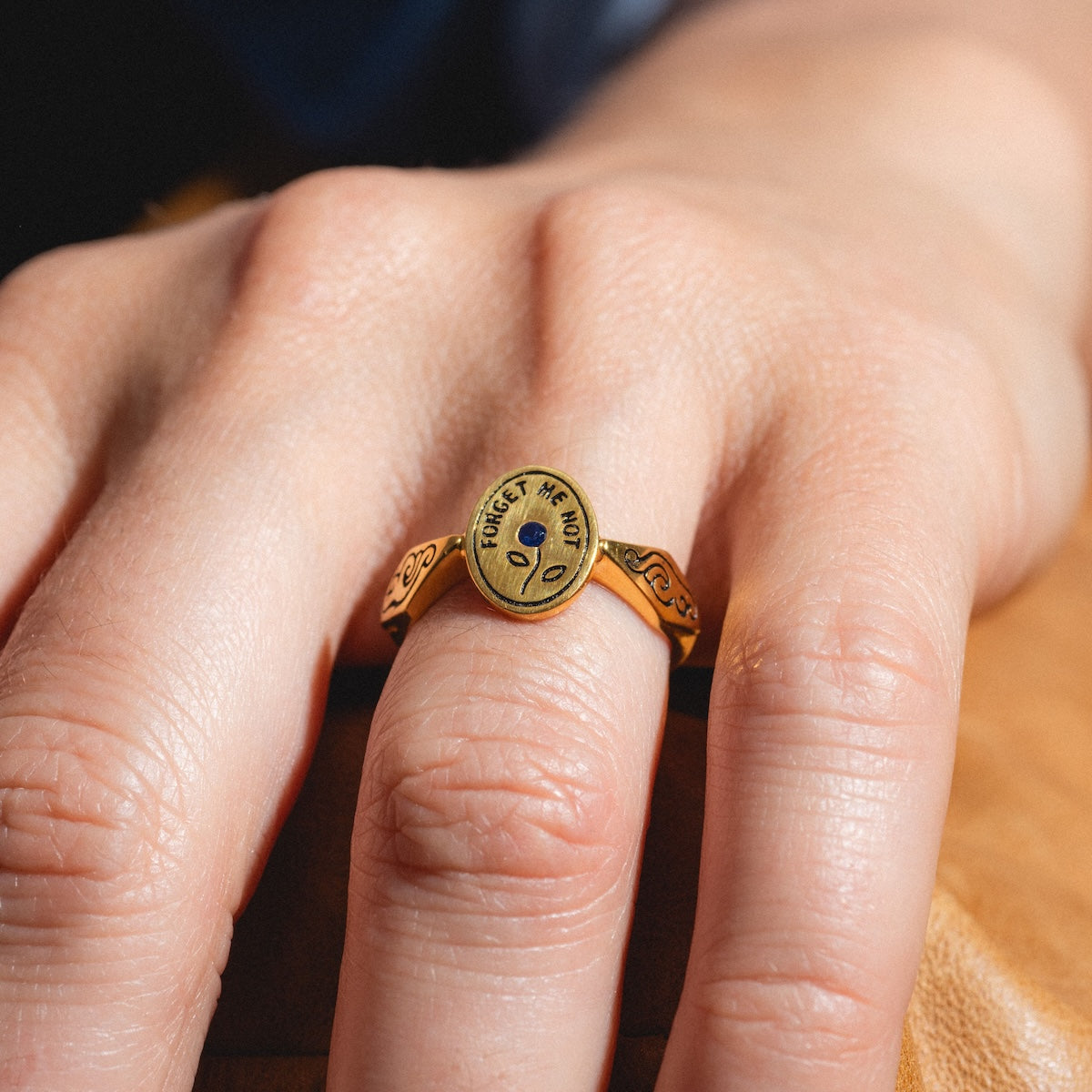 A close-up of a hand wearing the Forget Me Not Ring, a yellow gold vermeil signet ring with an oval face, engraved with FORGET ME NOT and featuring a small blue flower at the center.