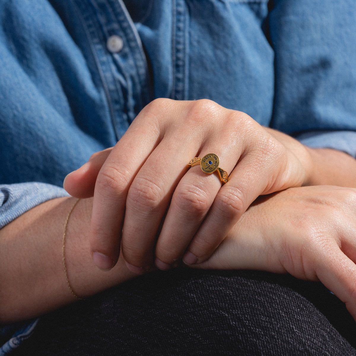 A person dressed in a blue denim shirt and thin gold bracelet rests their hands on their knee, wearing the Forget Me Not Ring—a yellow gold vermeil signet ring featuring a yellow smiley face design.