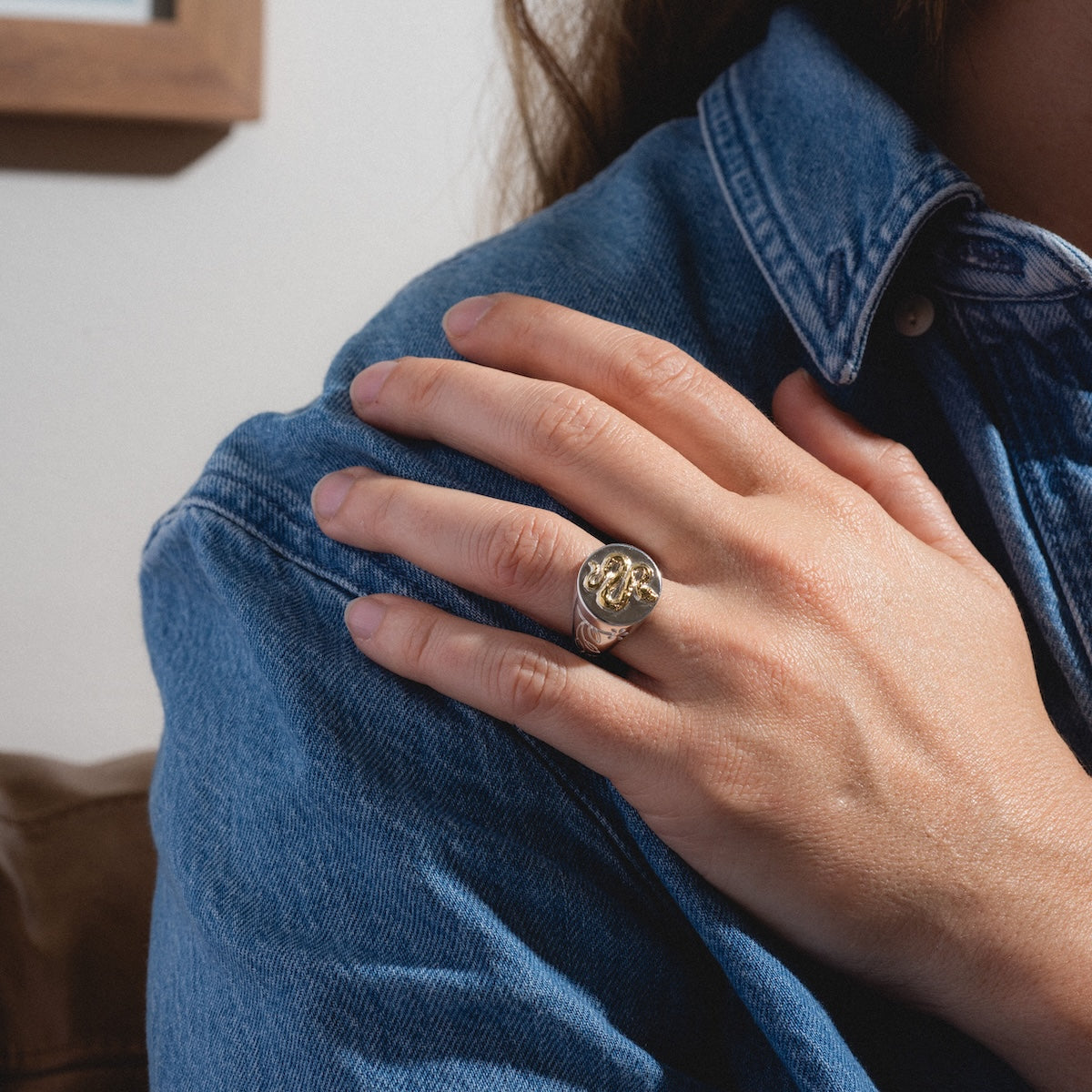 A person wearing a blue denim shirt rests their hand on their shoulder, displaying the Serpent Signet Ring—a gold ring with delicate floral engravings—on their finger.