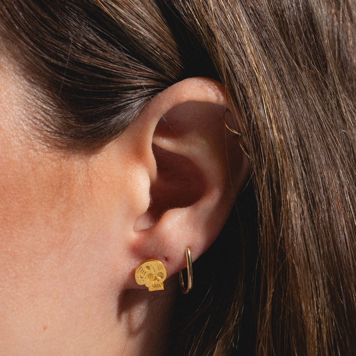 Close-up of a person’s ear with brown hair, wearing two gold earrings: a small round 18k gold Skull Stud Earring and a simple hoop on the earlobe.