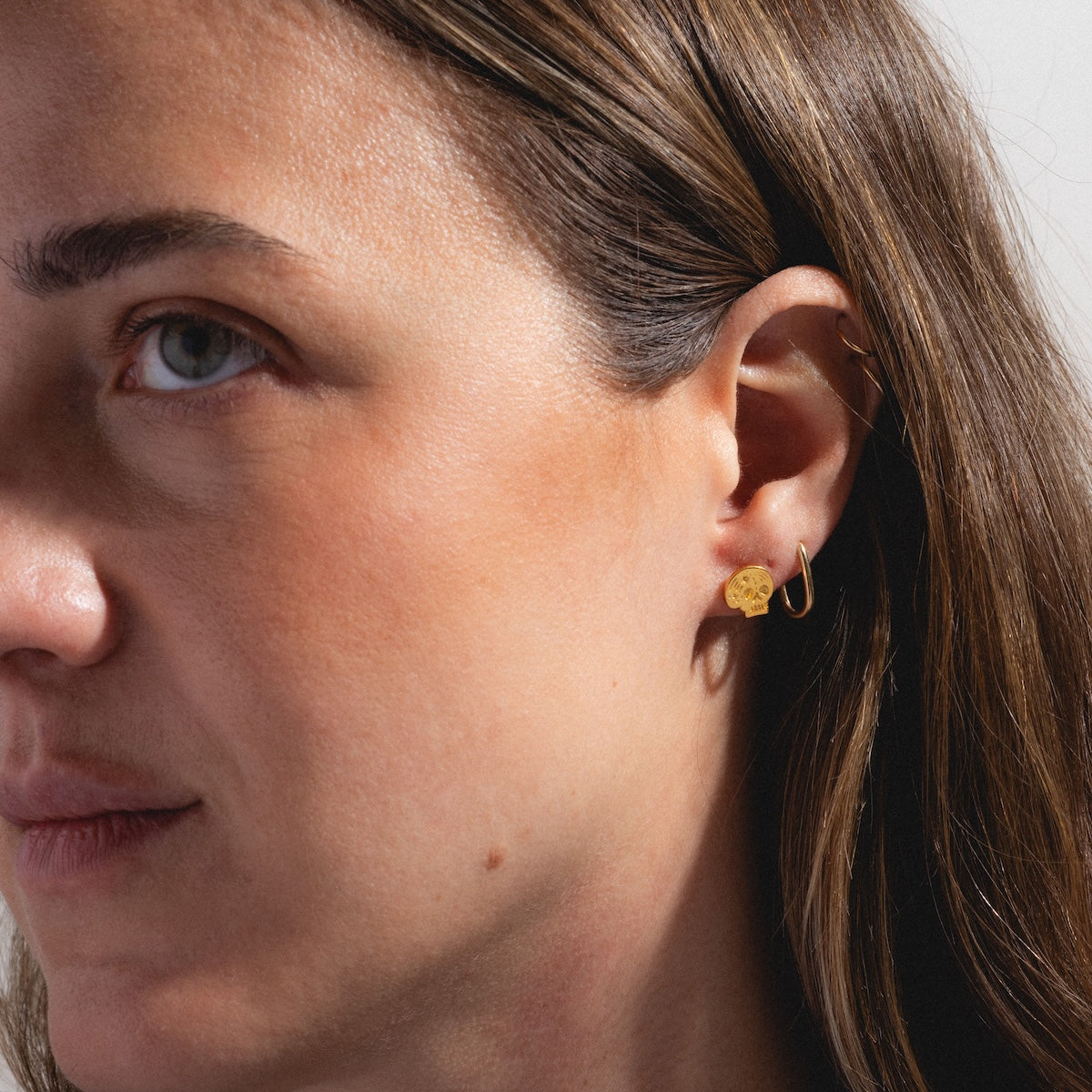 Close-up of a woman with light skin and brown hair, focusing on her ear adorned with Skull Stud Earrings in 18k gold and a minimalist gold hoop. The background is neutral.