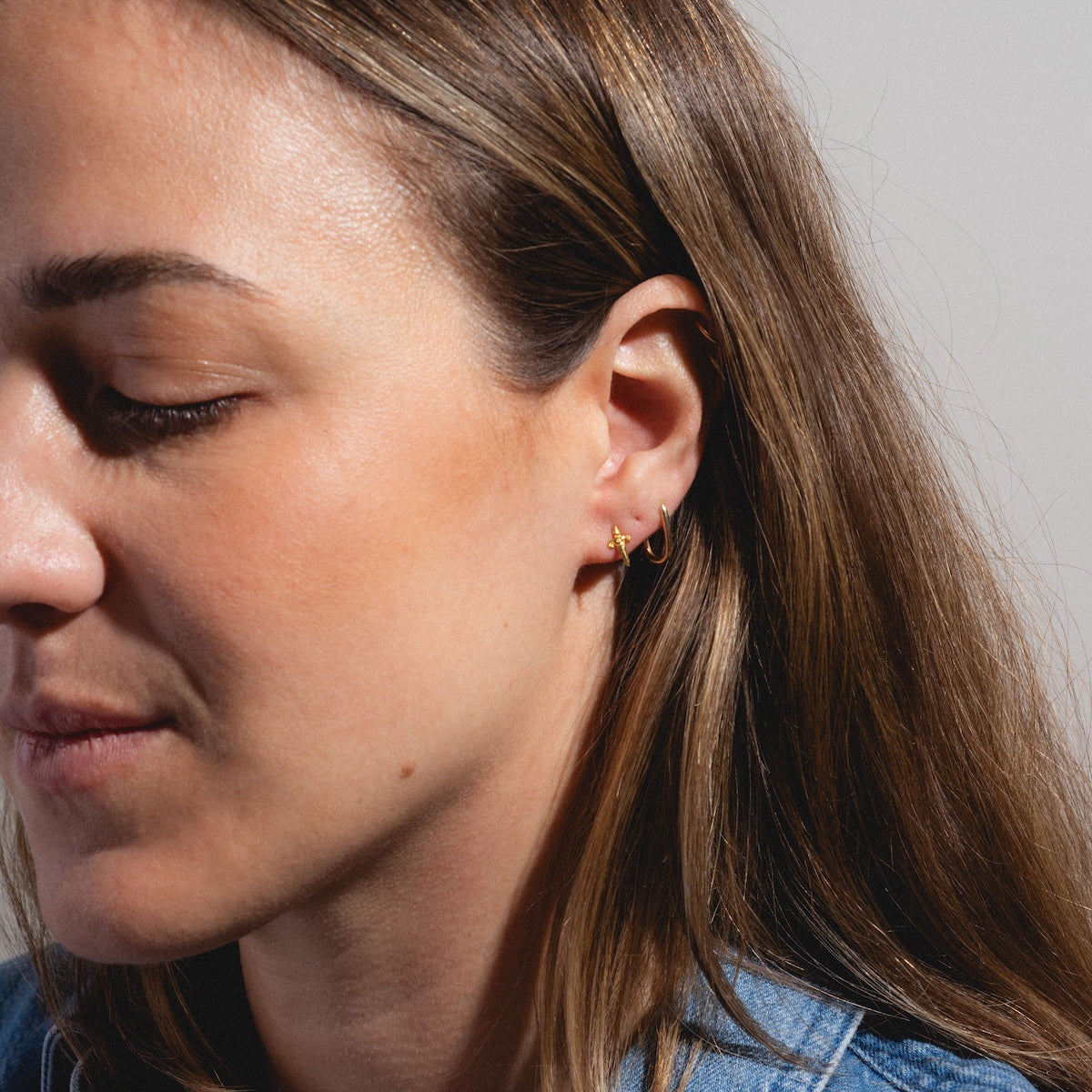 A close-up of a woman with long brown hair wearing two Mini Dagger Stud Earrings by Hunt of Hounds, looking down with a neutral expression against a plain background.