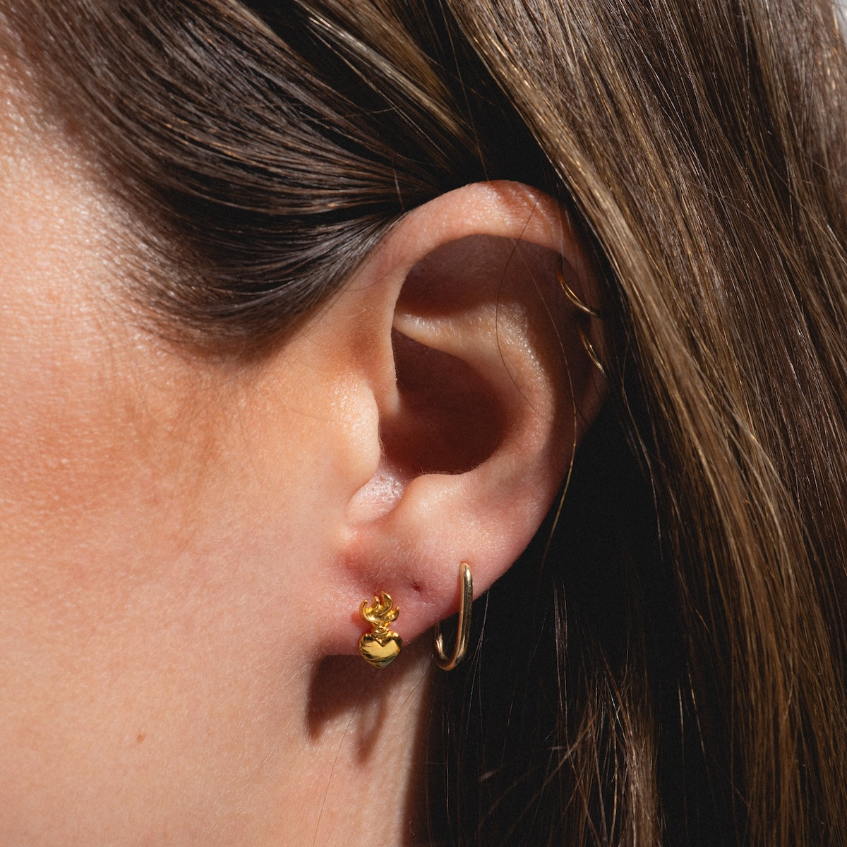 Close-up of a persons ear with two gold vermeil earrings: a small hoop in the lobe and the Sacred Heart Stud Earring above it. Brown hair partially covers the upper ear, highlighting the playful mix of stud and hoop earrings.
