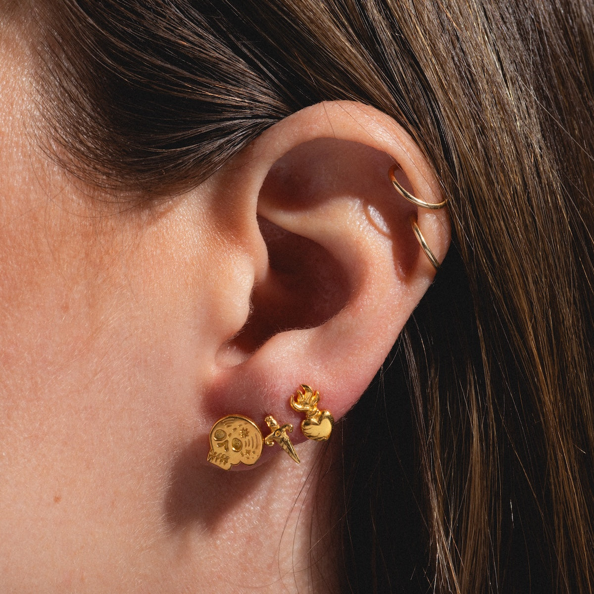 A close-up of a person’s ear with three gold vermeil stud earrings, including the Sacred Heart Stud Earrings, plus a gold hoop in the upper ear. The person has straight brown hair.