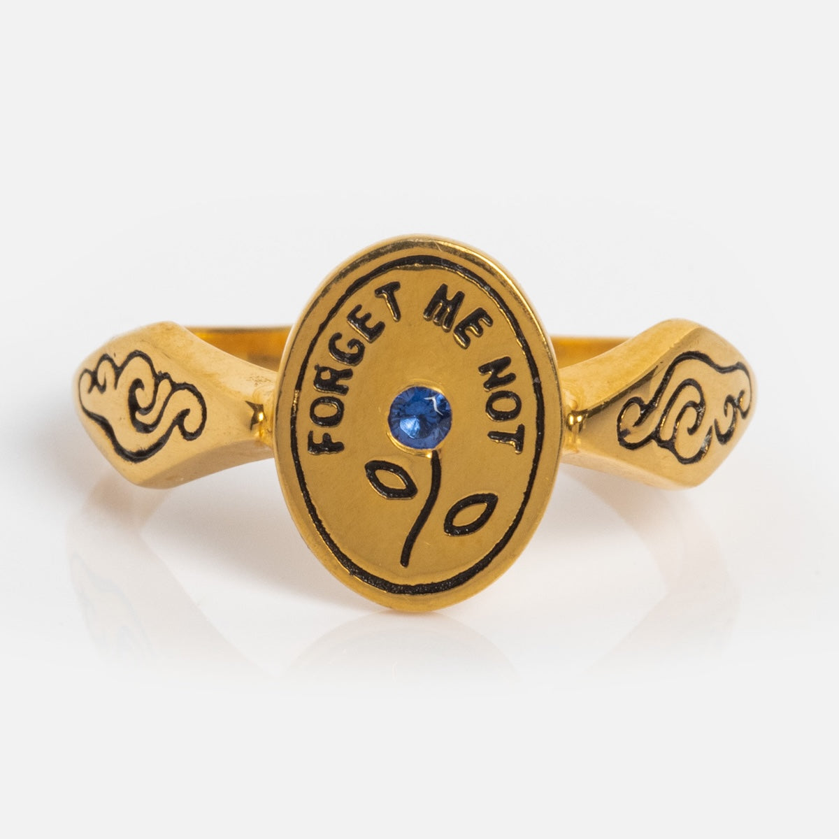 The Forget Me Not Ring is a yellow gold vermeil signet with an oval face engraved “FORGET ME NOT,” featuring a central blue gemstone, a simple sprout design, and a band adorned with black decorative patterns.