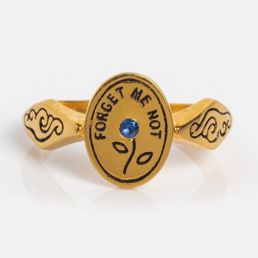 The Forget Me Not Ring is a yellow gold vermeil signet with an oval face engraved “FORGET ME NOT,” featuring a central blue gemstone, a simple sprout design, and a band adorned with black decorative patterns.