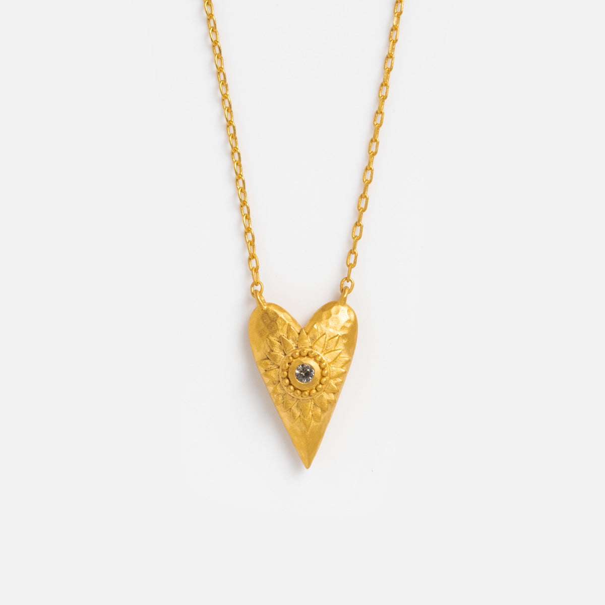 The I Am Loved Heart Necklace features a textured gold heart pendant with a sunburst design and a central white topaz stone, hanging from a delicate gold chain, displayed on a plain white background.